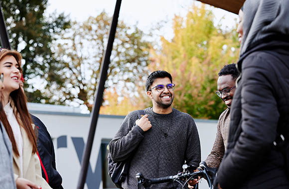 Four people standing outdoors, smiling and talking together near a fence with autumn trees in the background.