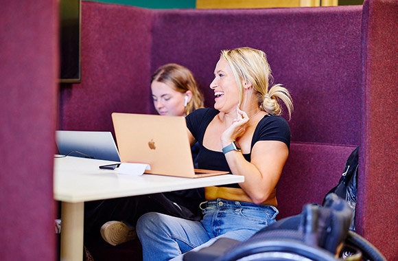 Two people sitting together in a booth, smiling and working on laptops.
