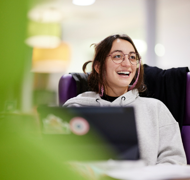 A student sits in the lounge working on her laptop and smiling.