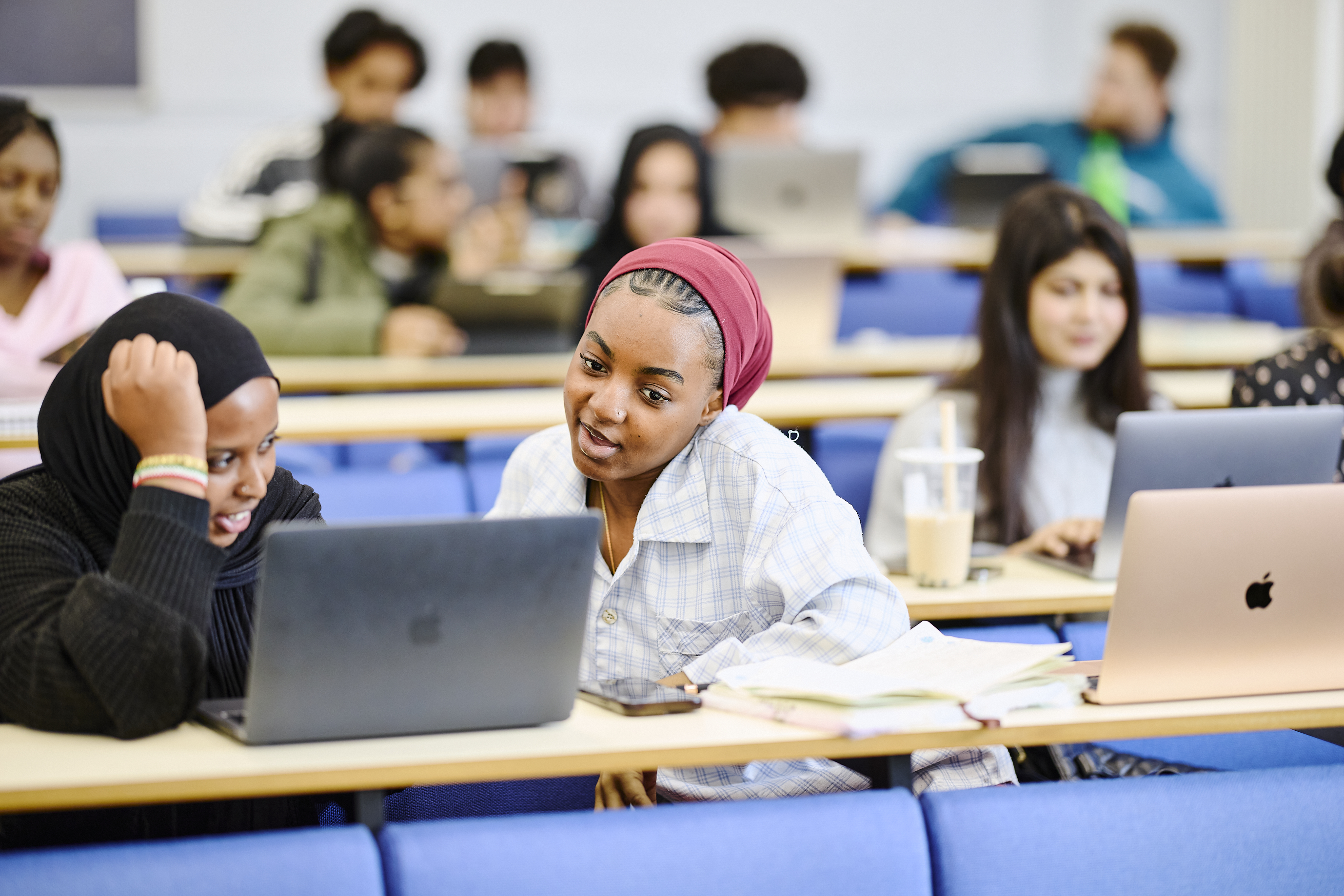 Two students sit in front of a computer, discussing a topic in a lecture theatre.