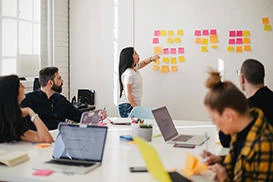 Business students sit in a meeting room as one student talks through post-it notes on a whiteboard.