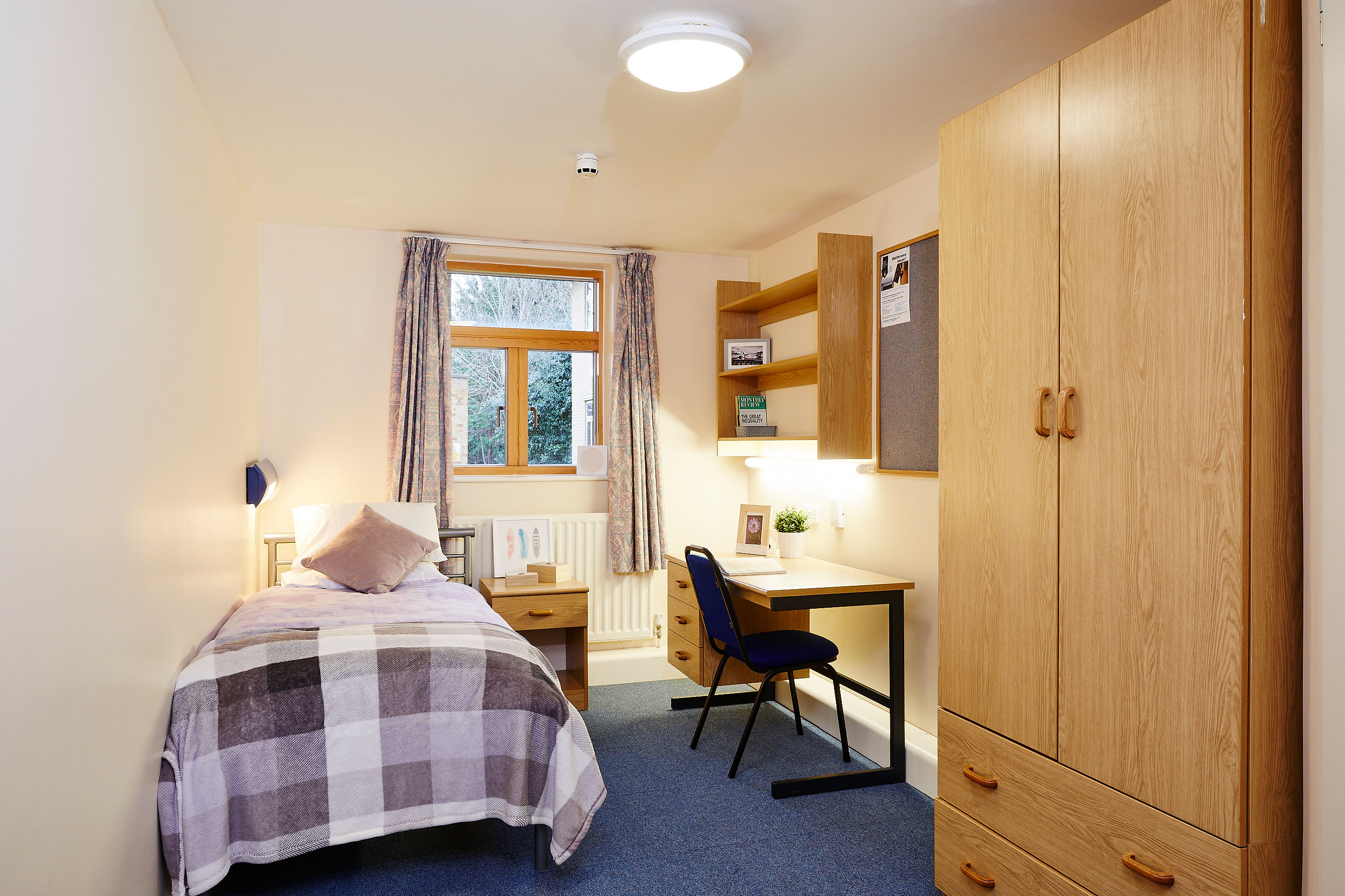 Single student bedroom with a bed, wooden wardrobe, desk and chair by a window.