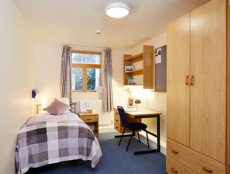 Single student bedroom with a bed, wooden wardrobe, desk and chair by a window.