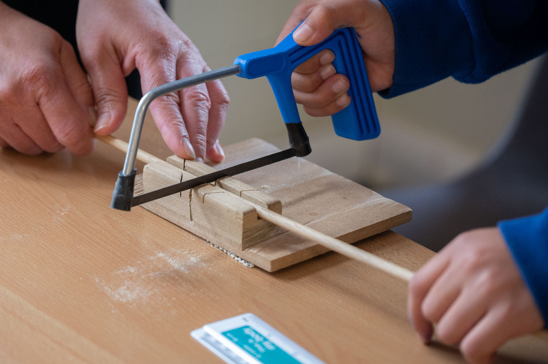 Two people sawing a wooden dowel on a table with a blue-handled saw, using a wooden guide for accuracy.