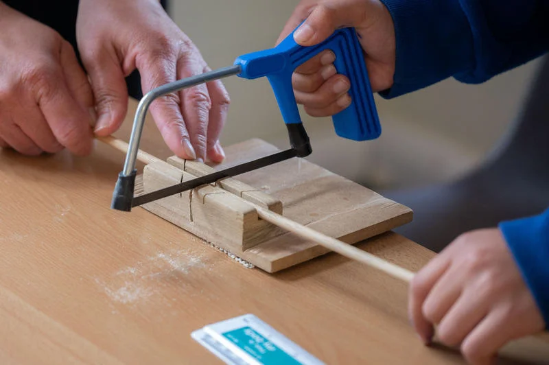 Two people sawing a wooden dowel on a table with a blue-handled saw, using a wooden guide for accuracy.