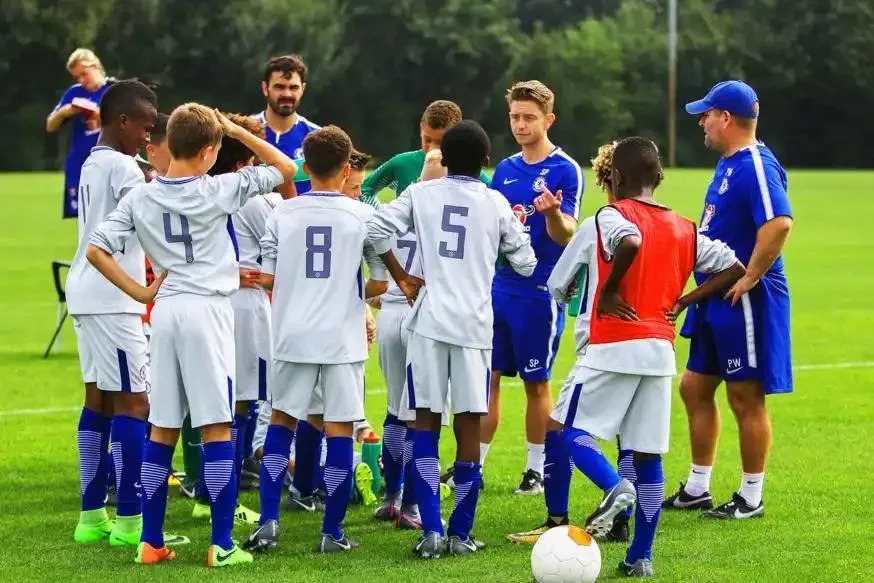 A football coach stands talking with his team on the pitch before a game.