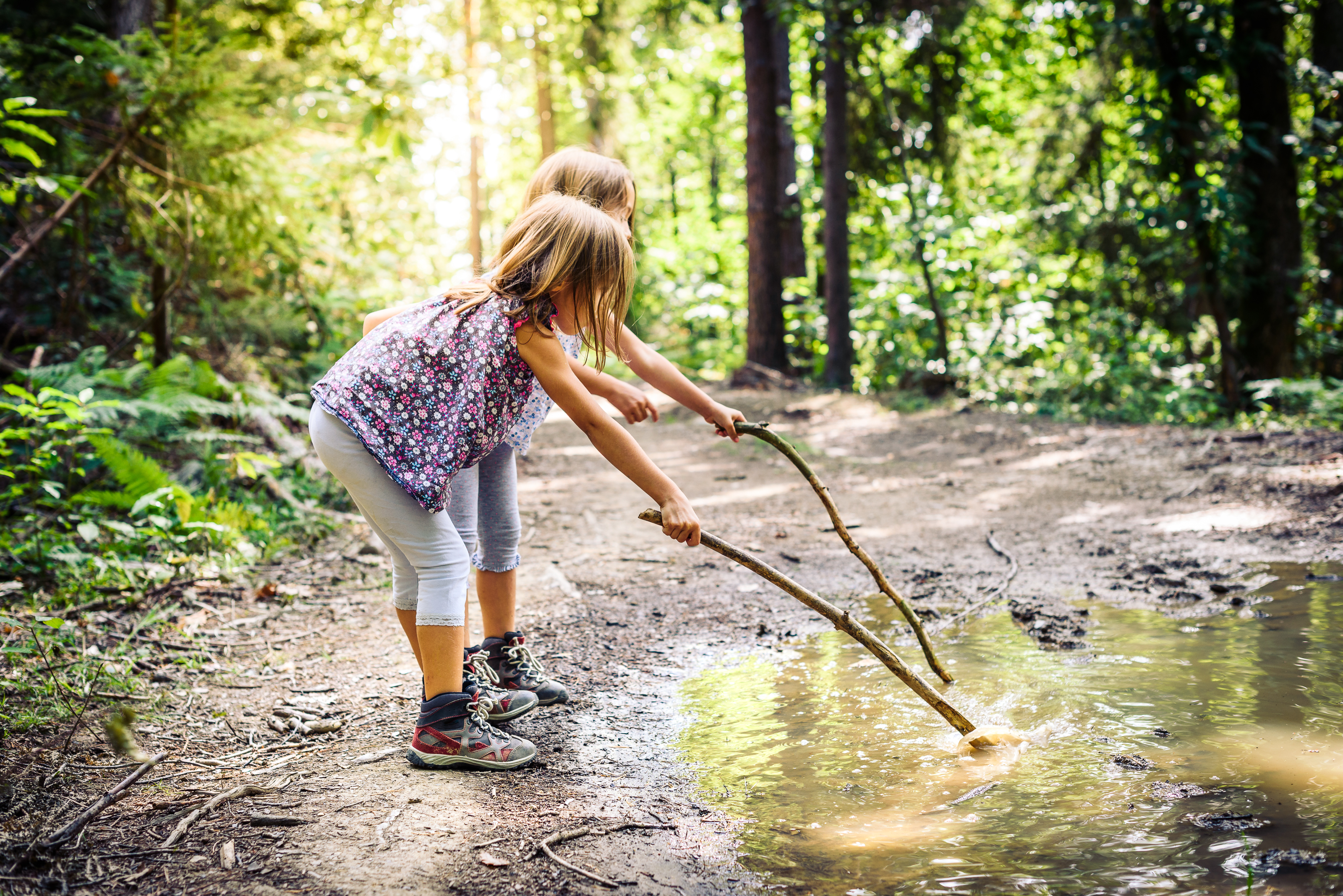 2 children playing with sticks in a puddle in woodland.
