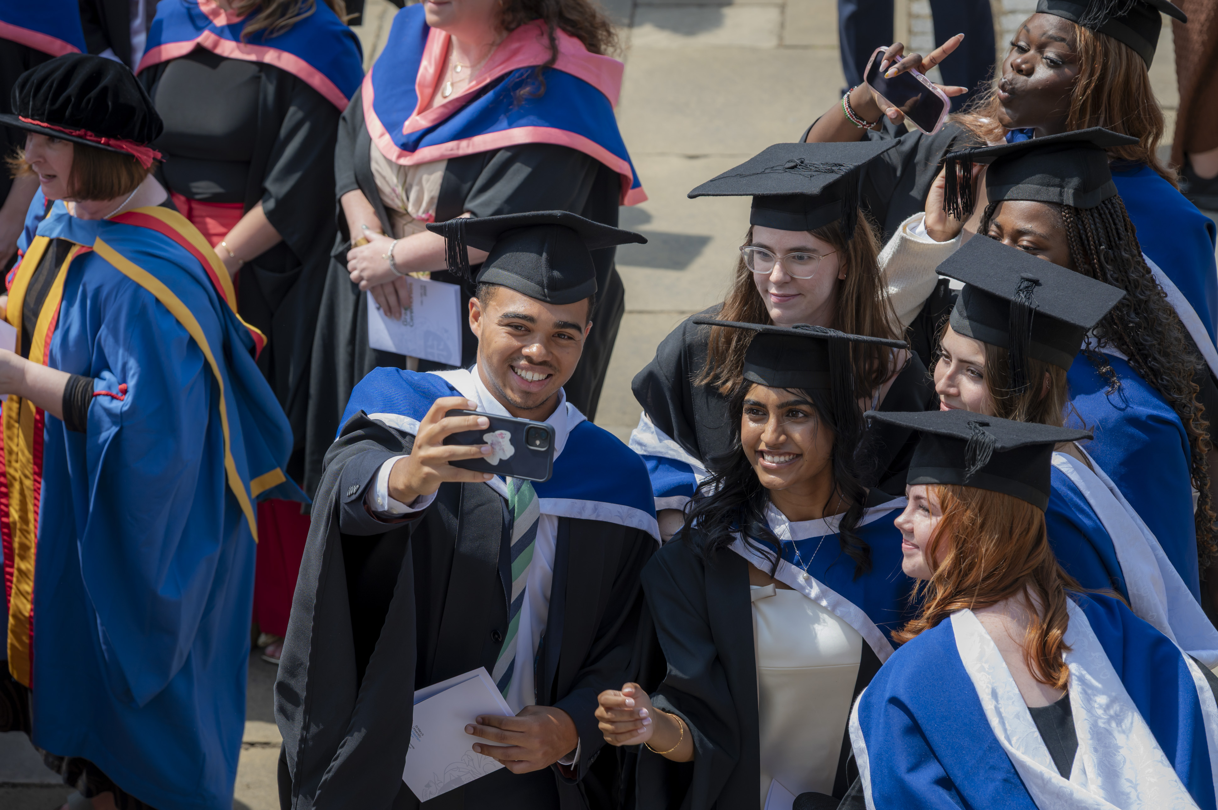 Graduates in caps and gowns smile and take a selfie together at a graduation ceremony.