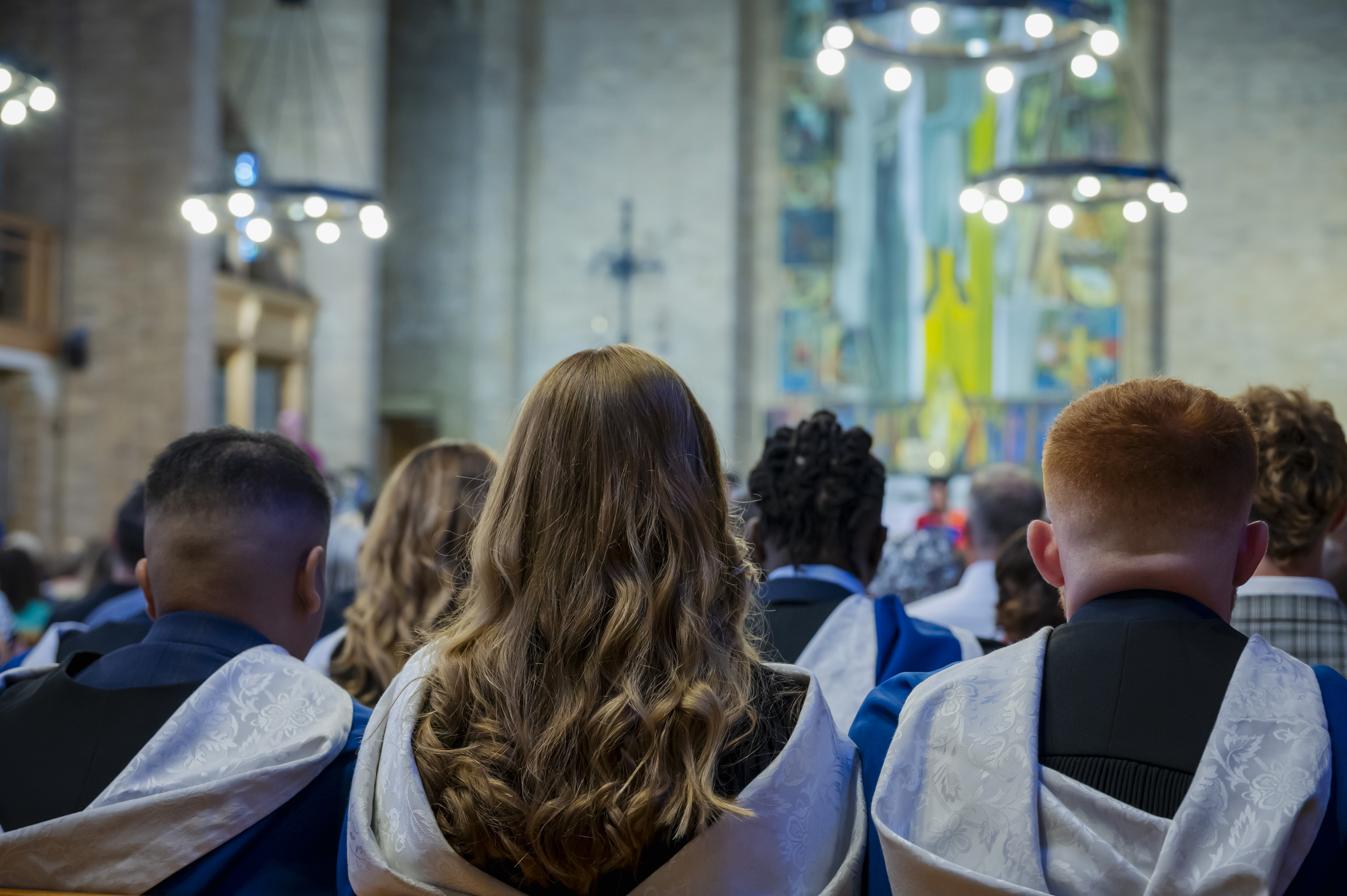 Graduates in caps and gowns sit facing a stage or altar in a decorated hall or church during a ceremony.