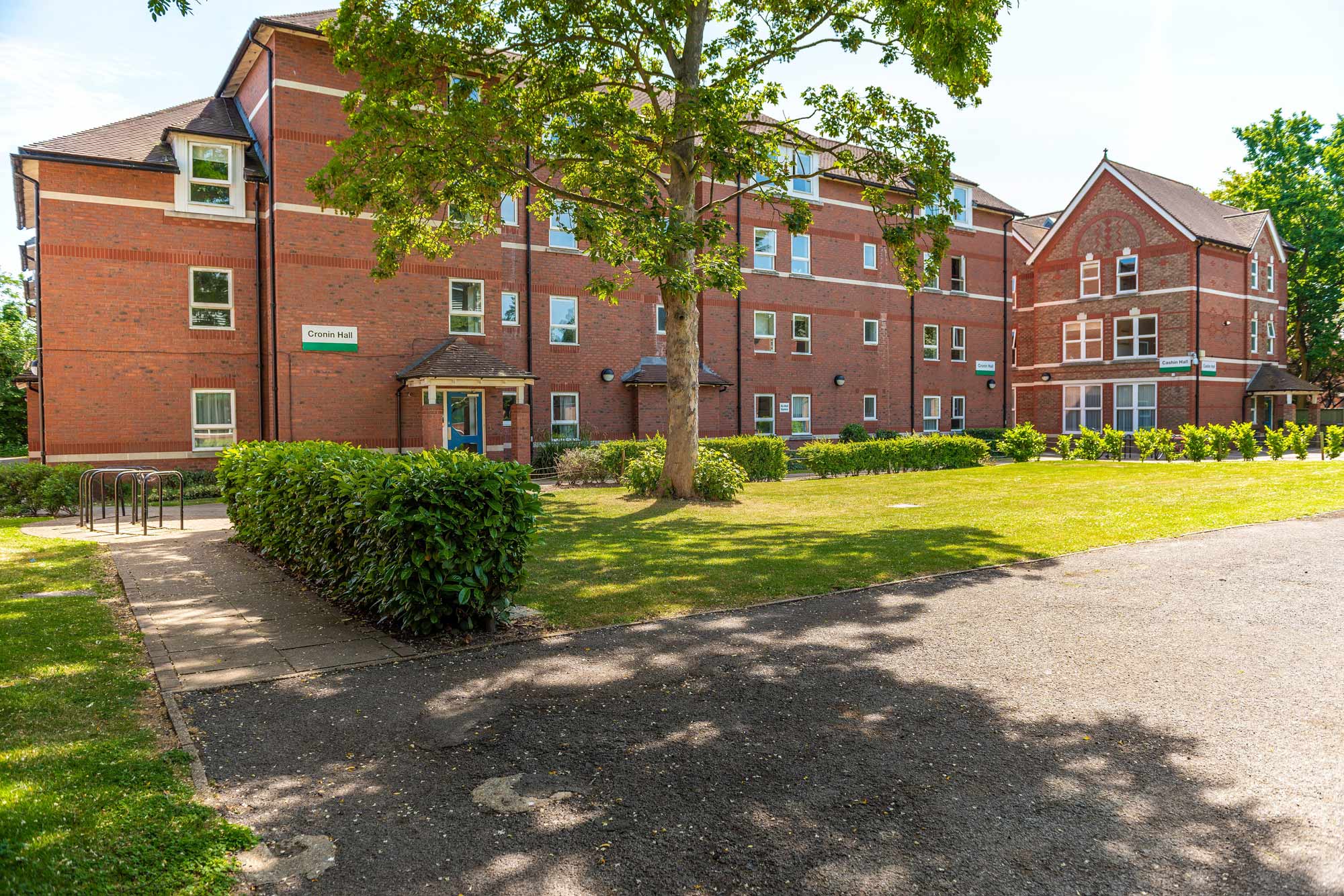 Red brick apartment building with multiple windows, surrounded by green lawns, trees, and a paved walkway in front.
