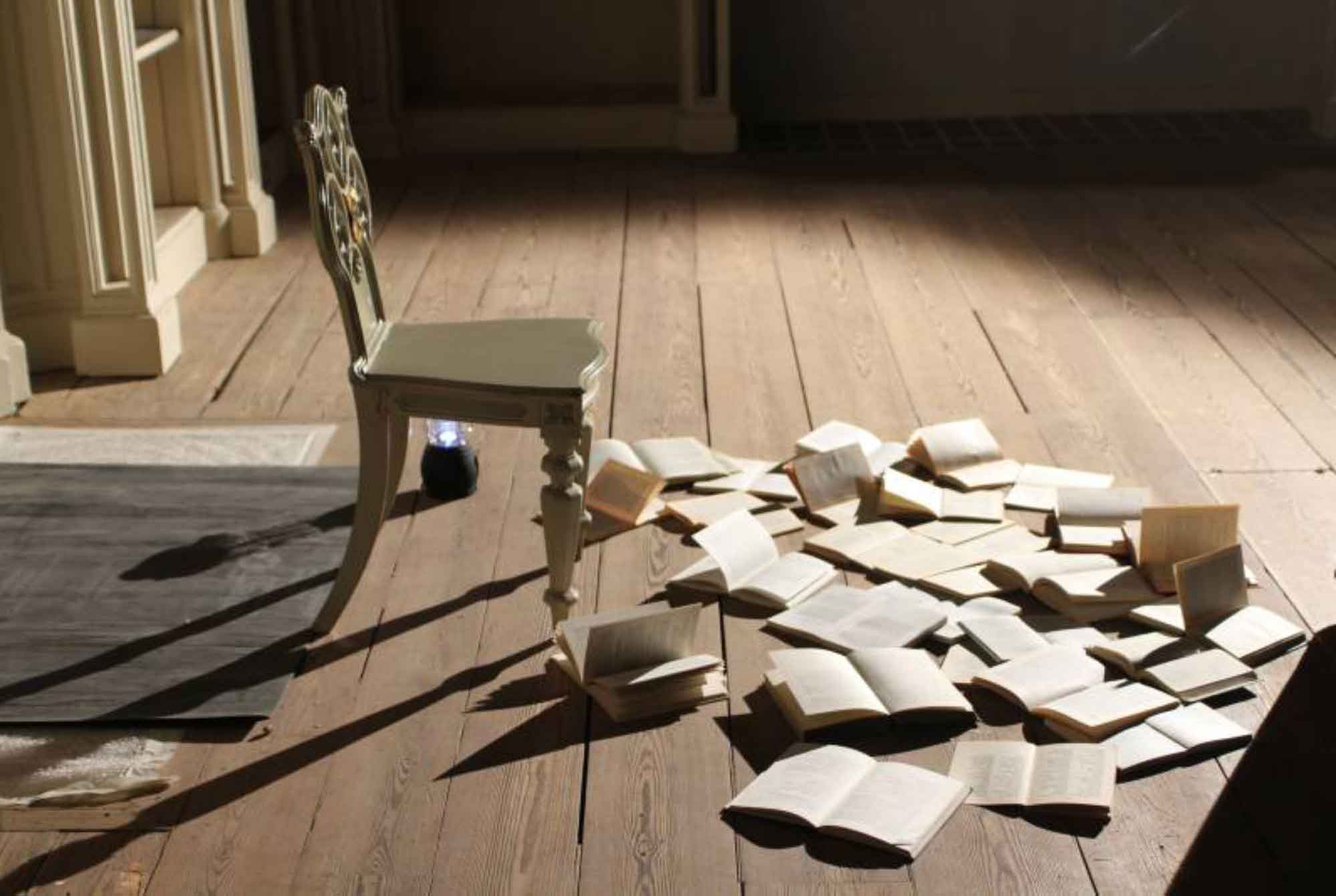 A small chair and a pile of open books scattered on a wooden floor in a sunlit room.