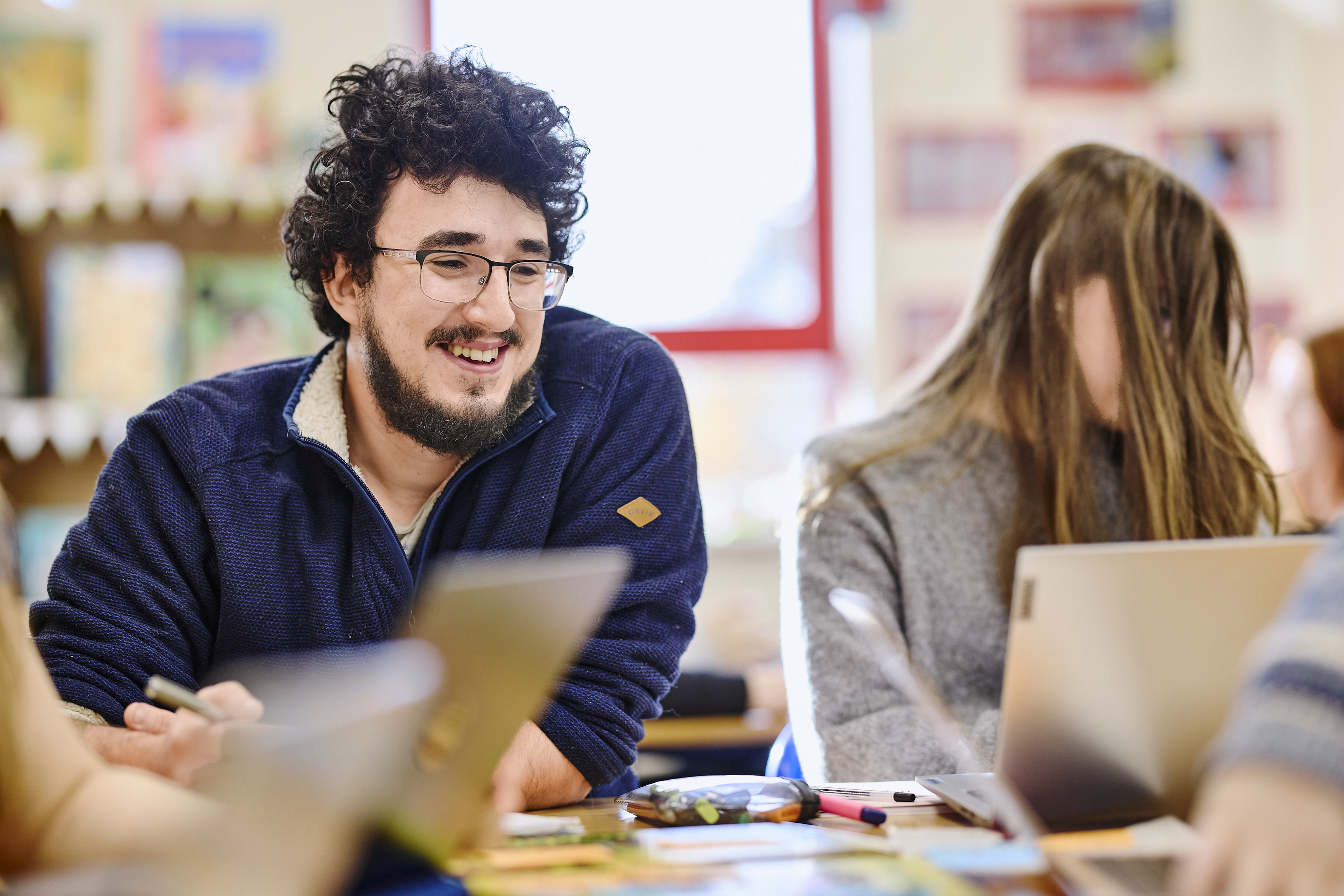 Two mature students sitting at a table, one smiling and the other writing, with books and papers in a classroom setting.