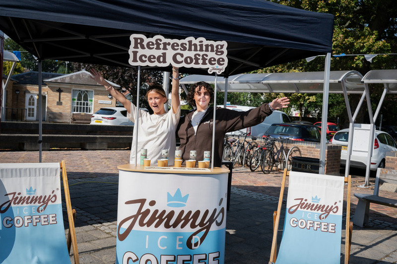 Two people smiling at an outdoor booth labelled "Jimmy's" holding a sign that reads "Refreshing Iced Coffee".