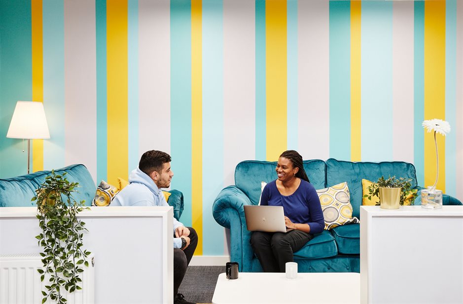 Two people sit and talk on blue velvet sofas. One of them has a laptop. 