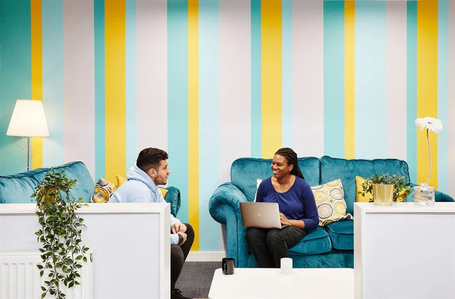 Two people sit and talk on blue velvet sofas. One of them has a laptop.