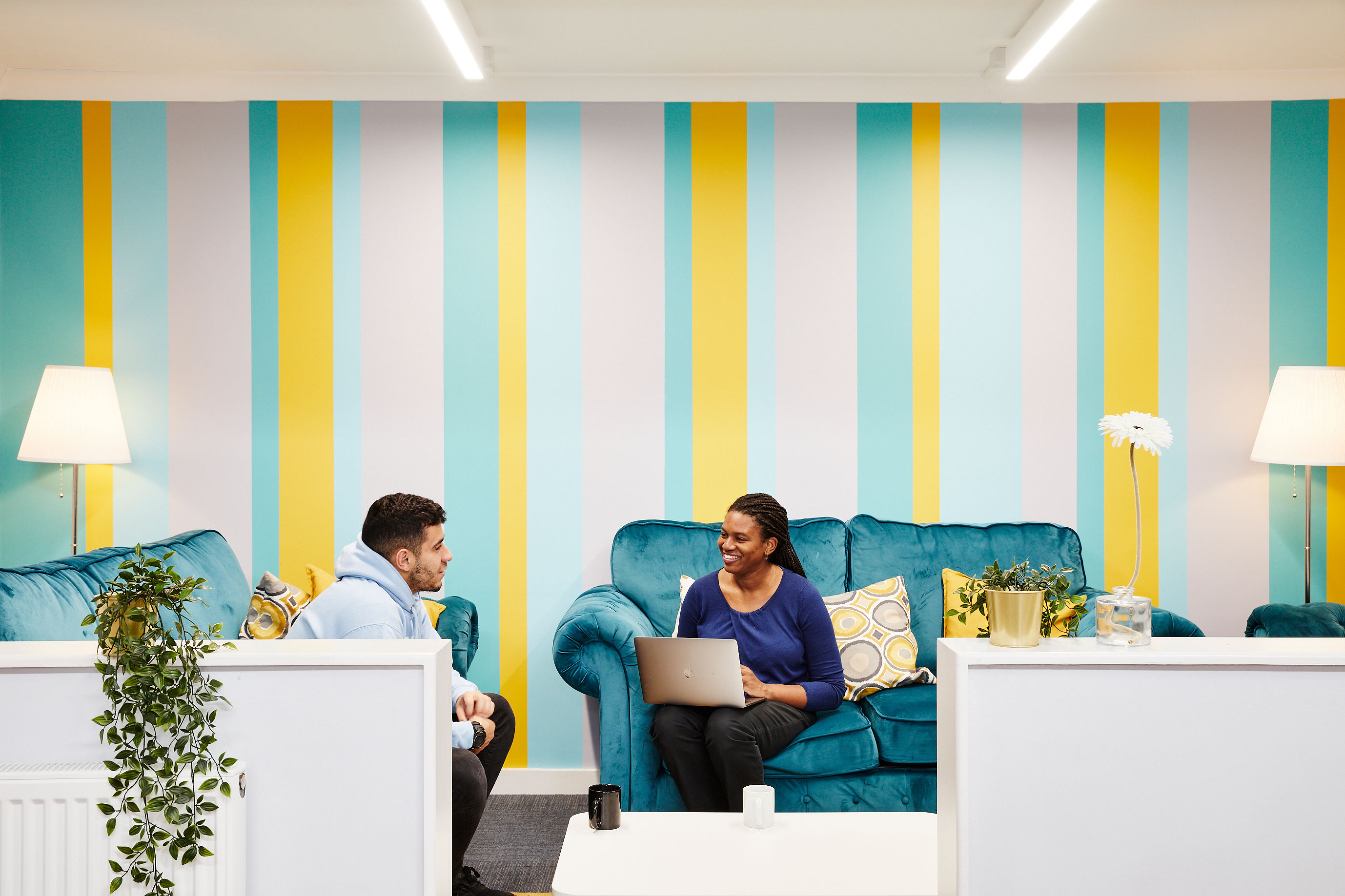 Two people sit and talk on blue velvet sofas. One of them has a laptop. 