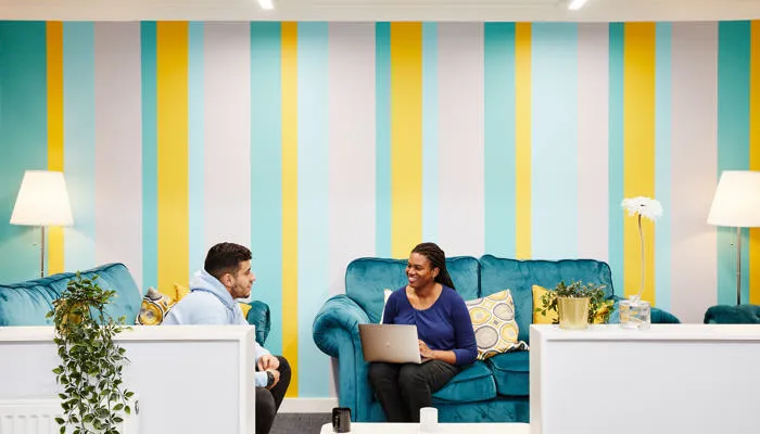 Two people sit and talk on blue velvet sofas. One of them has a laptop.
