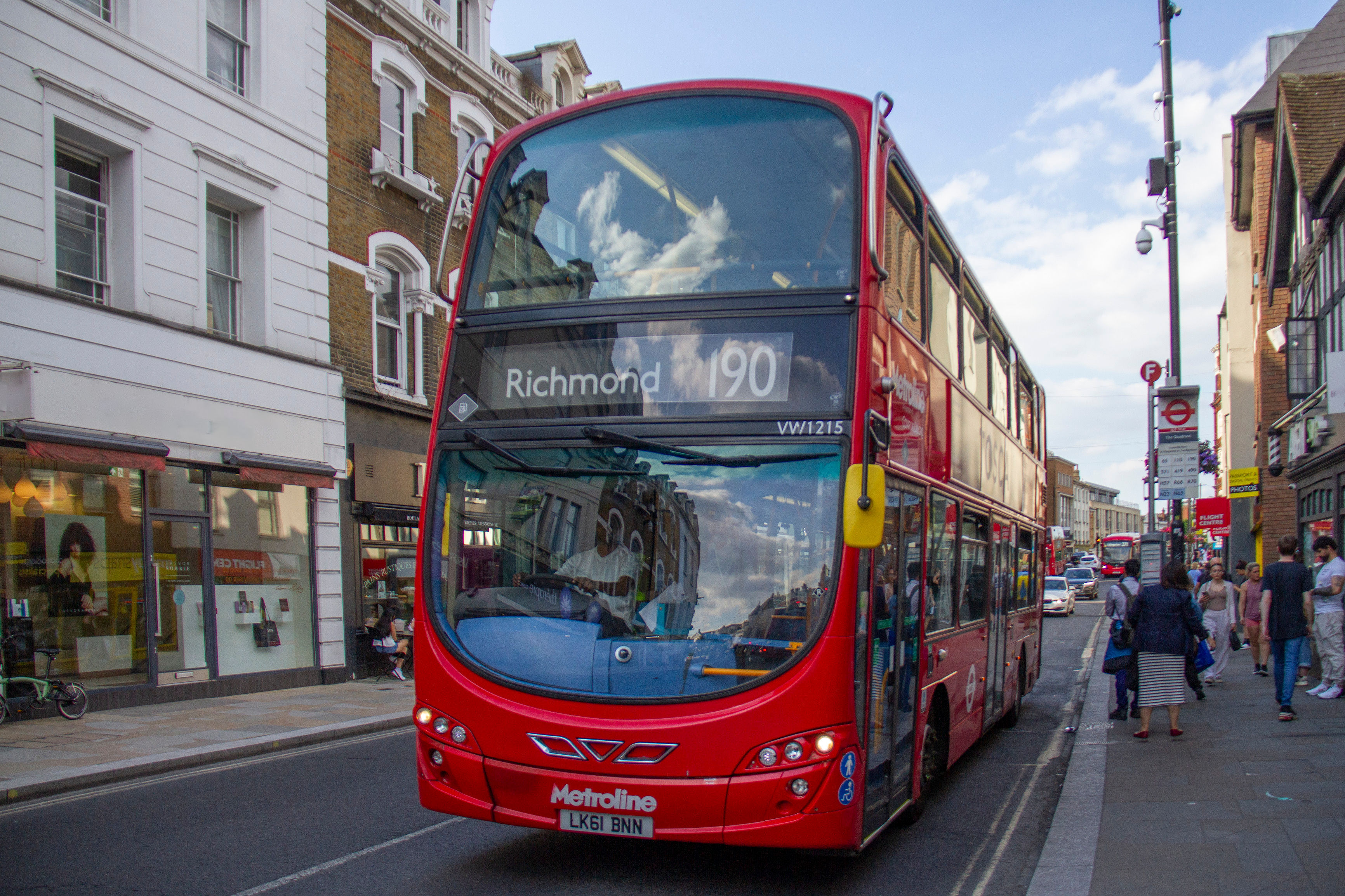 Red double-decker bus driving on a city street with shops and pedestrians on either side.