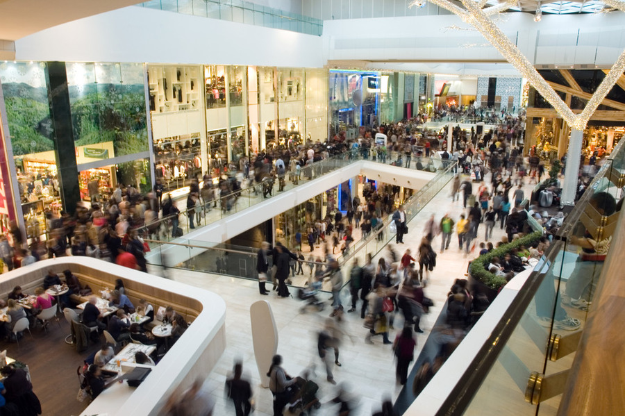 Crowded shopping centre with multiple levels, stores, and people walking and gathering throughout the space.