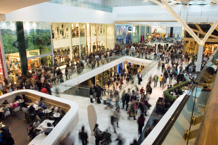 Crowded shopping centre with multiple levels, stores, and people walking and gathering throughout the space.