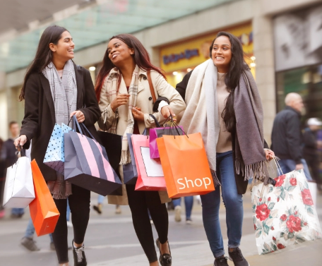 Three women walking outdoors, smiling and carrying multiple shopping bags.