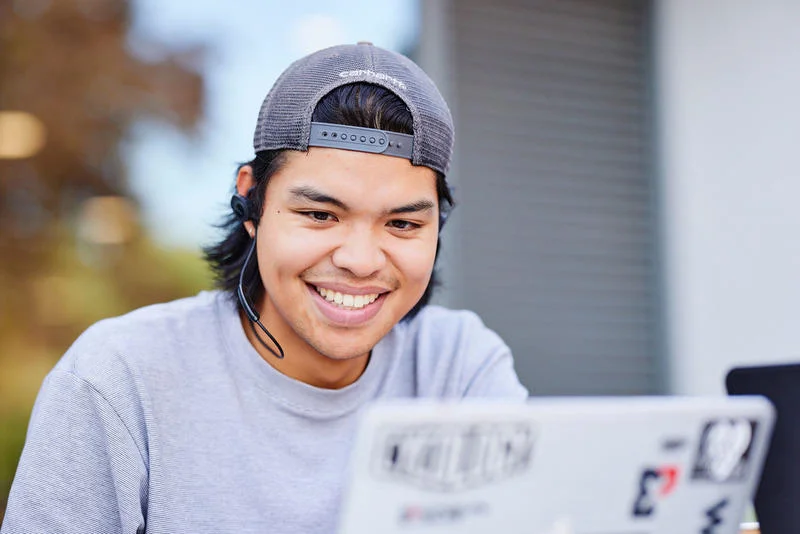 Smiling person wearing a backwards cap using a laptop with stickers, sitting outdoors.