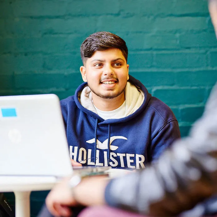 Smiling person in a blue hoodie sitting at a table with a laptop, talking to another person.