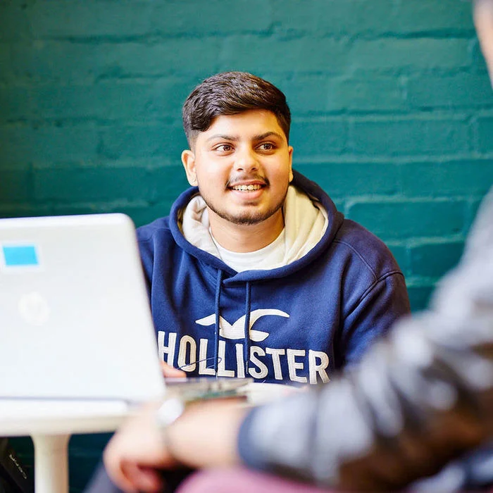 Smiling person in a blue hoodie sitting at a table with a laptop, talking to another person.