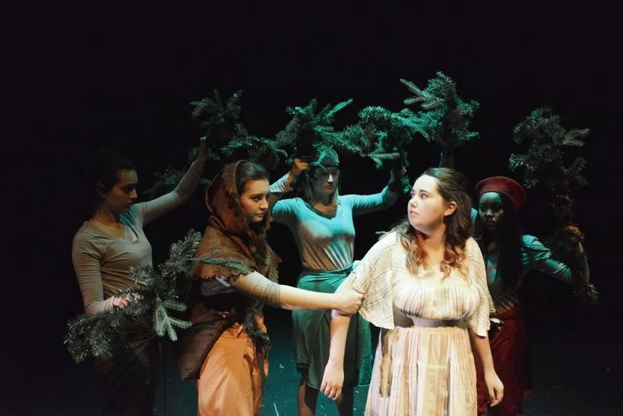 Actors in costume perform a dramatic scene on stage with a dark, leafy background.