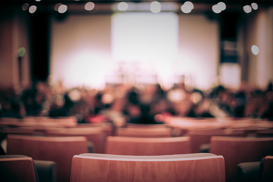 Rows of theatre seats in the foreground, with a blurred audience facing a brightly lit stage and screen.