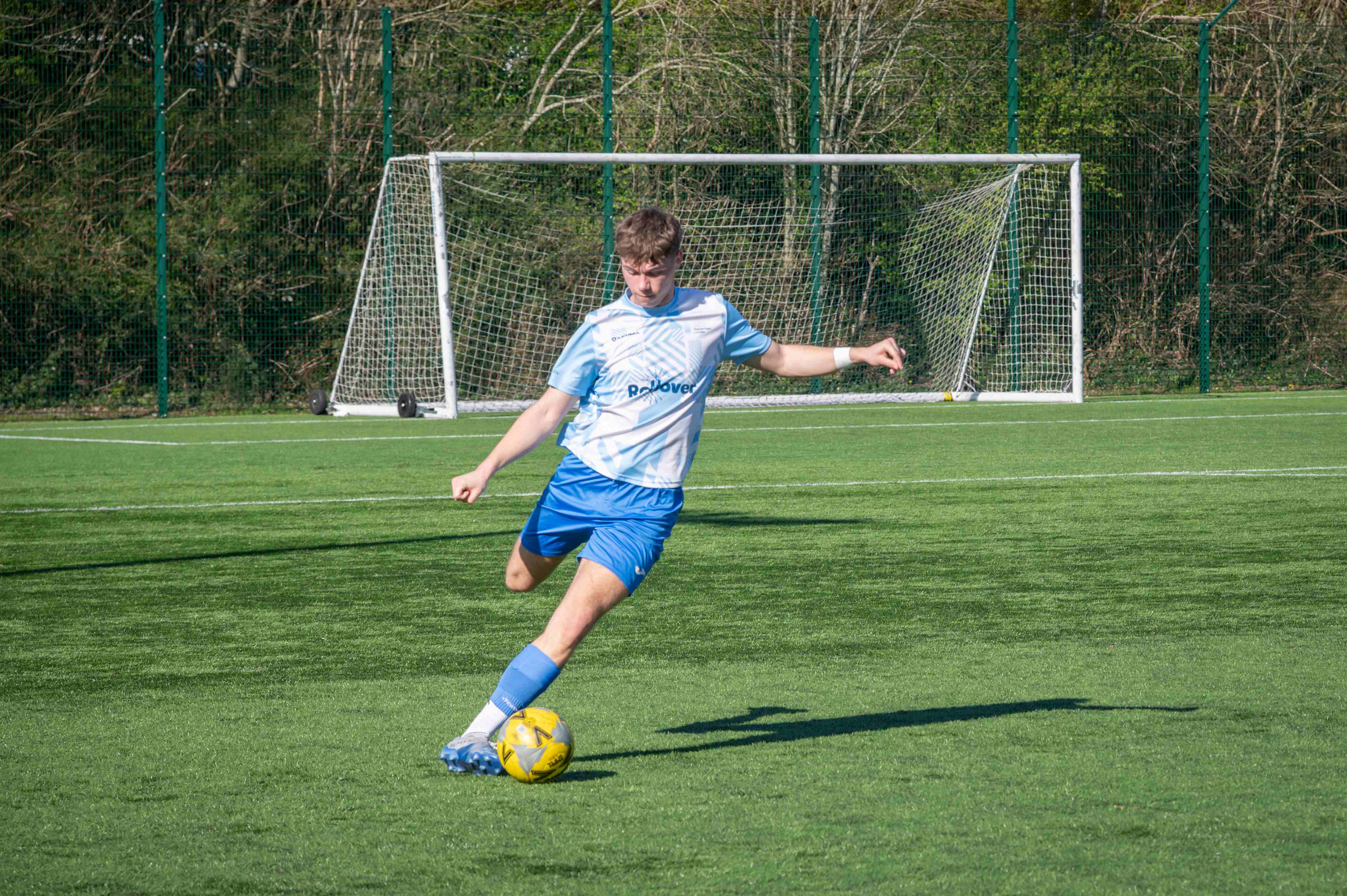 A footballer in a light blue and white kit prepares to strike a yellow football on an artificial grass pitch.