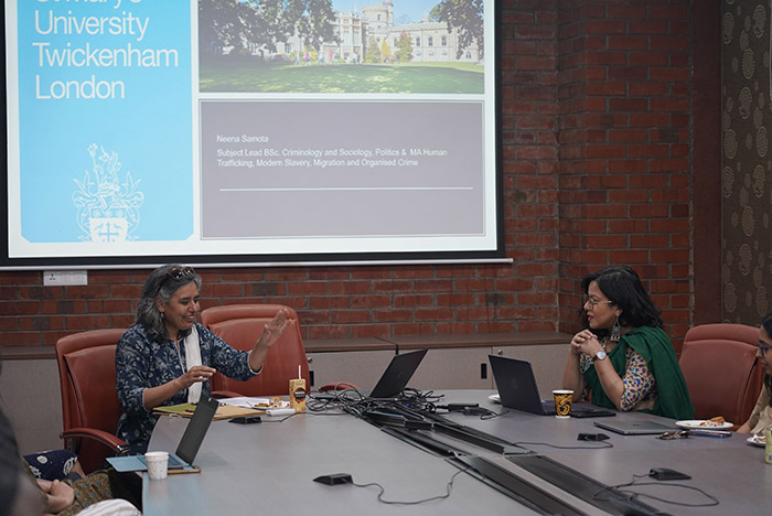 Neena Samota presenting whilst seated at board table with large screen behind her