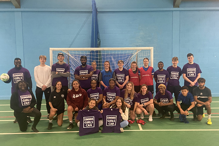 group of students standing in front of indoor football goal holding Simmie Girls Can t-shirts