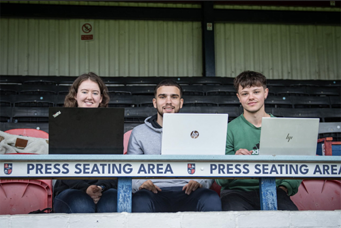 Three students sat in press seating area at football ground wiht laptops