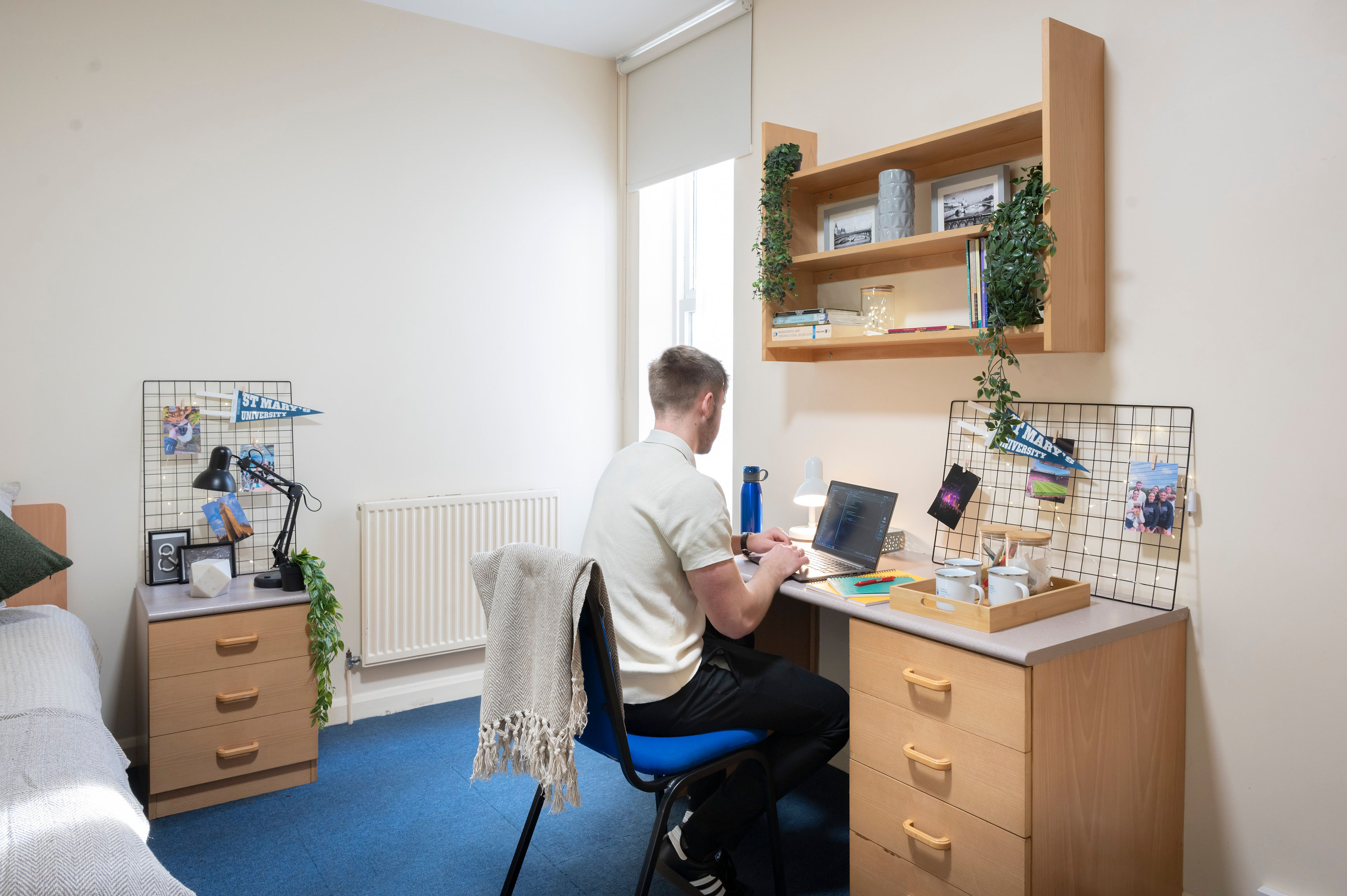 Person sitting at a desk in a tidy, well-lit dorm room, with books, photos, and plants on shelves.