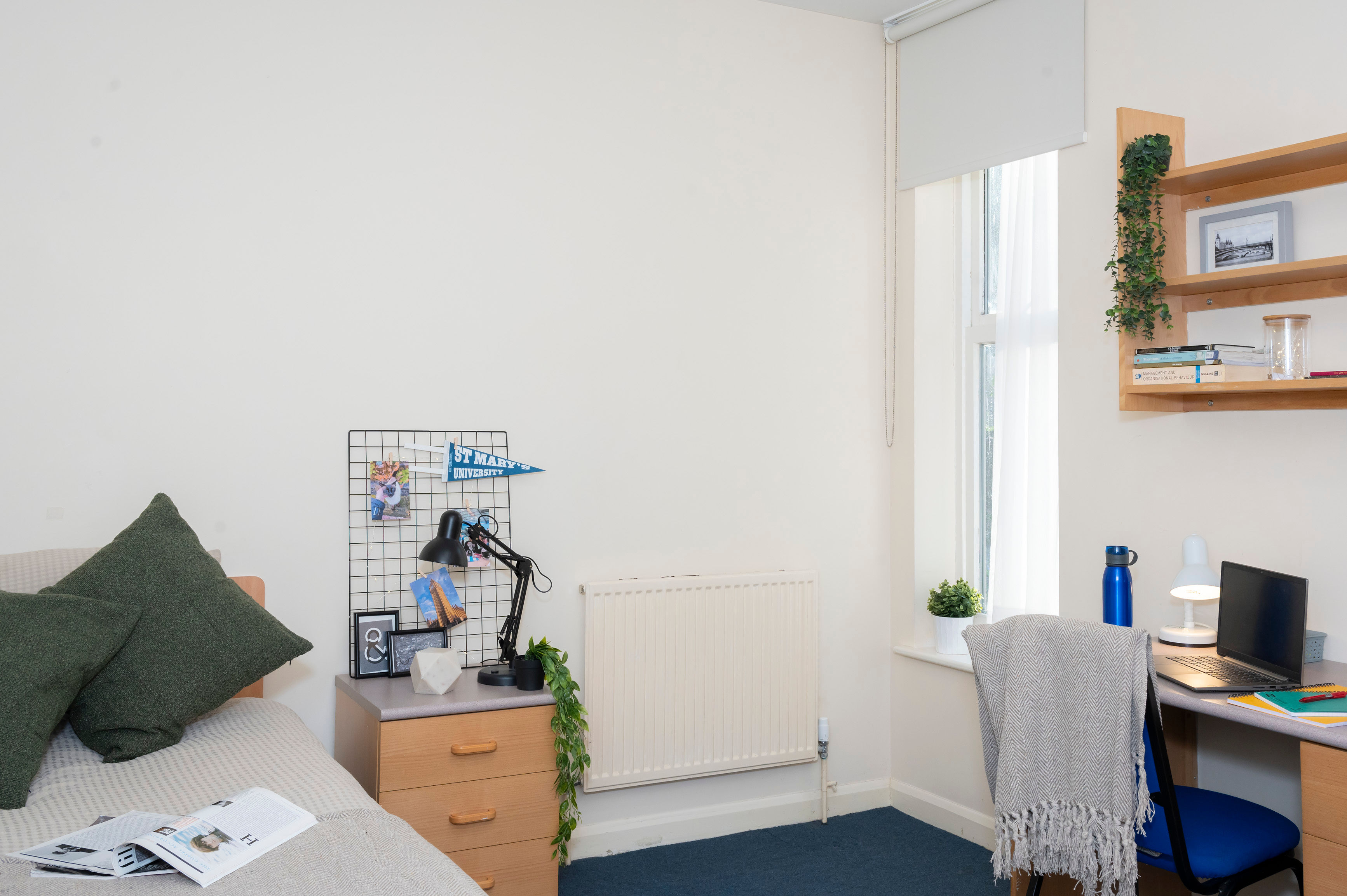 Small tidy bedroom with a bed, desk, shelves, and a window letting in natural light.