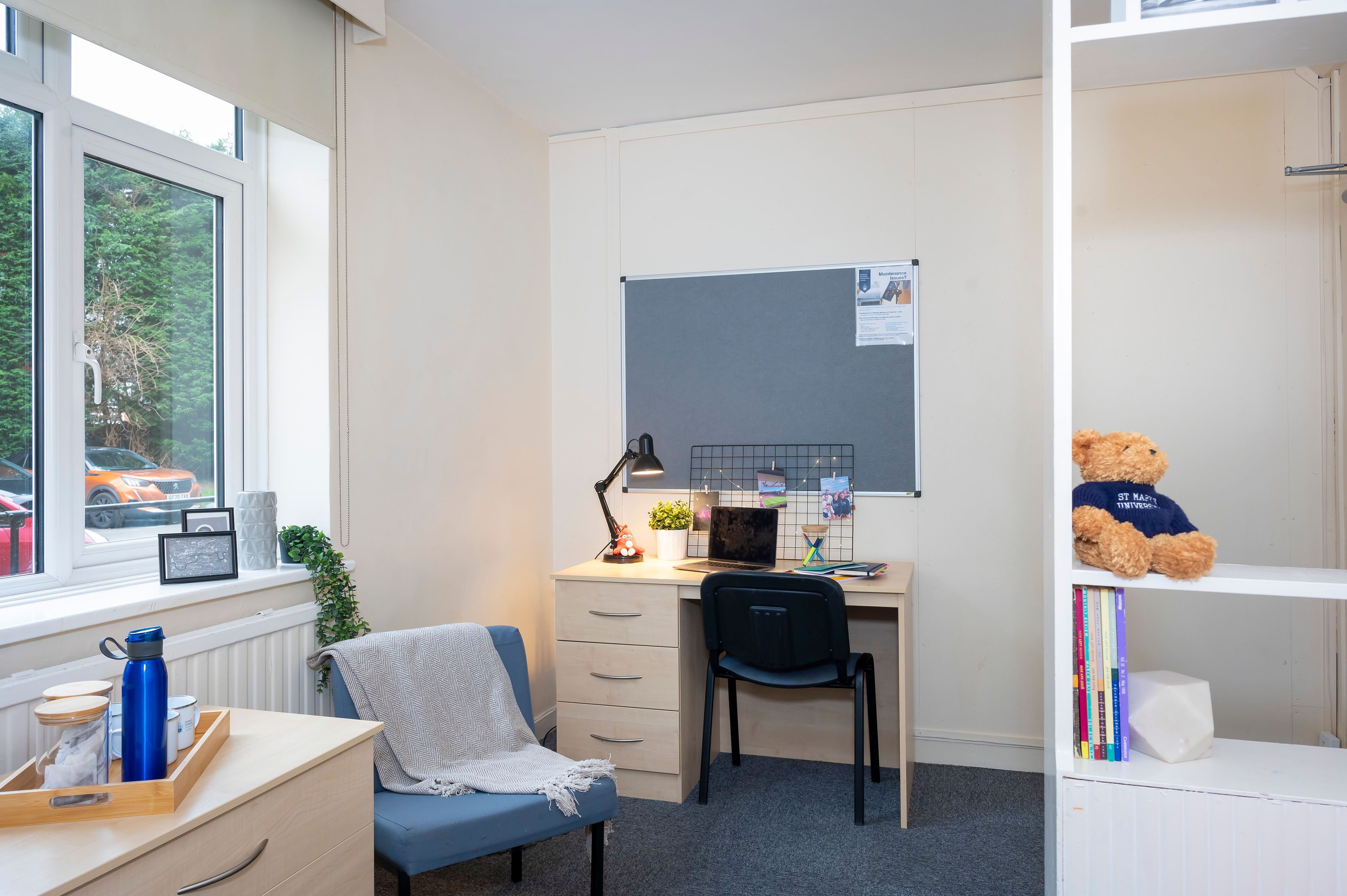 Neatly organized student dorm room with a desk, chair, bed, bulletin board, and shelves with a teddy bear.