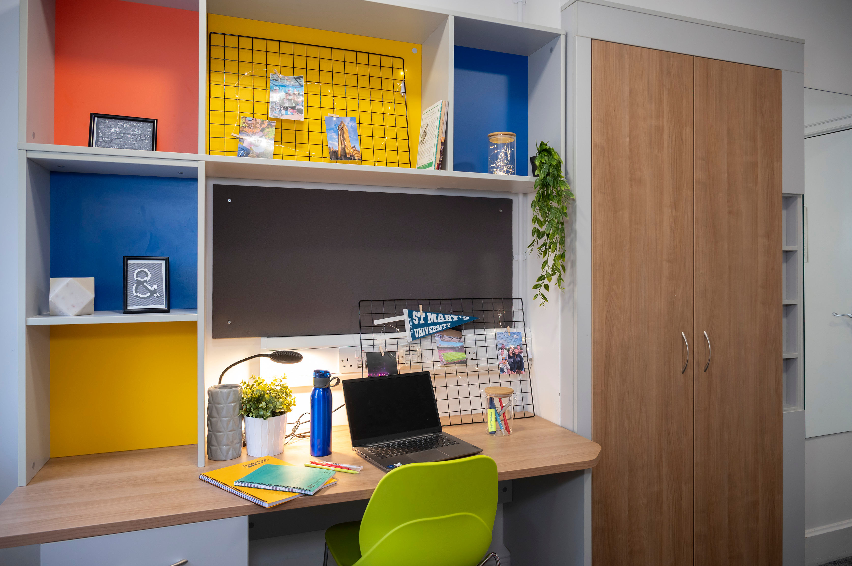 Desk with a laptop, stationery, and books, surrounded by colorful shelves and a green chair in a tidy study space.